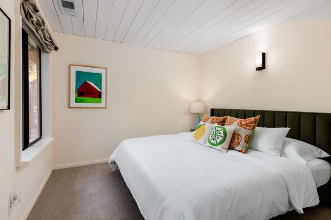 Cozy modern bedroom with large white bed, olive-green upholstered headboard, citrus-print throw pillows, framed colorful barn artwork, bedside lamp, wall sconce and window