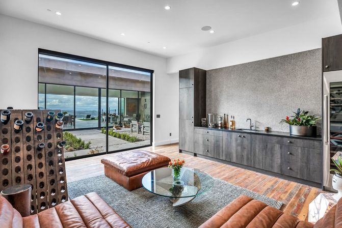 Bright modern living room with leather seating, glass coffee table, wine rack and dark wood wet bar, sliding glass doors opening to a patio and distant landscape view.
