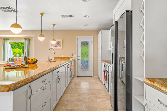 Sunny modern kitchen with long granite island, white shaker cabinets, globe pendant lights, stainless refrigerator and glass door opening to a green backyard.