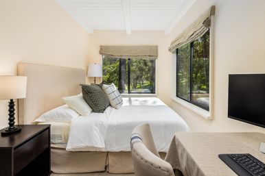 Bright neutral bedroom with white bedding and upholstered headboard, green-pattern pillows, bedside lamp on a dark nightstand, large corner windows with Roman shades looking out to leafy trees, and a small desk with monitor and keyboard — cozy work-from-home retreat.
