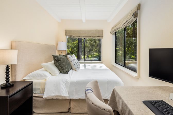 Bright neutral bedroom with white bedding and upholstered headboard, green-pattern pillows, bedside lamp on a dark nightstand, large corner windows with Roman shades looking out to leafy trees, and a small desk with monitor and keyboard — cozy work-from-home retreat.