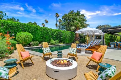 Sunny backyard pool patio with palm trees, a flaming round fire pit surrounded by wooden lounge chairs with colorful pillows, striped chaise lounges under a white umbrella and lush green hedges.