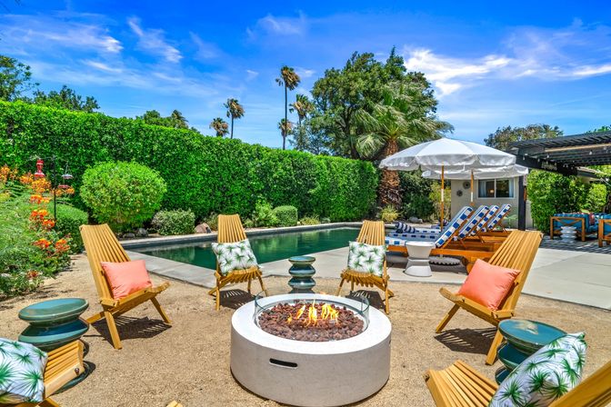 Sunny backyard pool patio with palm trees, a flaming round fire pit surrounded by wooden lounge chairs with colorful pillows, striped chaise lounges under a white umbrella and lush green hedges.