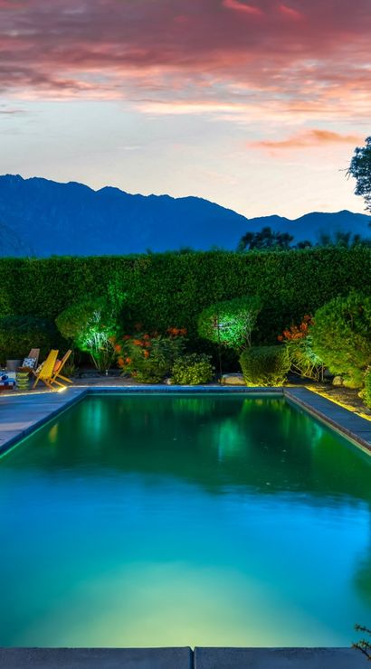 Dusk backyard with illuminated rectangular pool, striped lounge chairs and umbrellas on a patio, manicured hedges with landscape lighting, and silhouetted mountains under a pink sunset.
