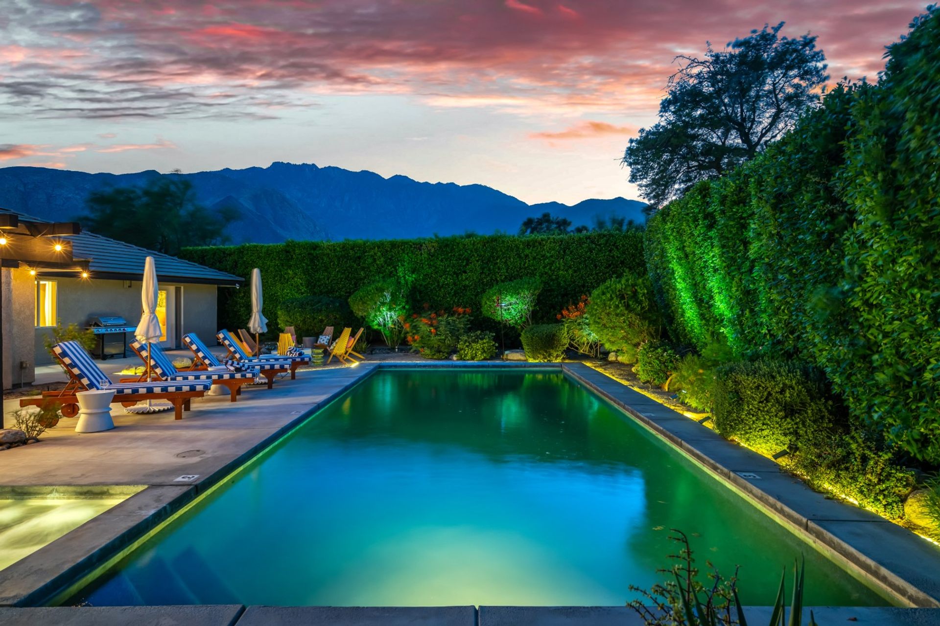 Dusk backyard with illuminated rectangular pool, striped lounge chairs and umbrellas on a patio, manicured hedges with landscape lighting, and silhouetted mountains under a pink sunset.