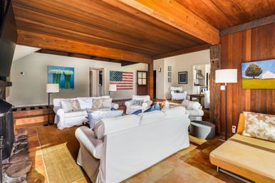Cozy sunlit living room with exposed wood-beamed ceiling, white slipcovered sofas, stone fireplace, wood-paneled wall, American flag wall art and colorful paintings on warm tile floors.