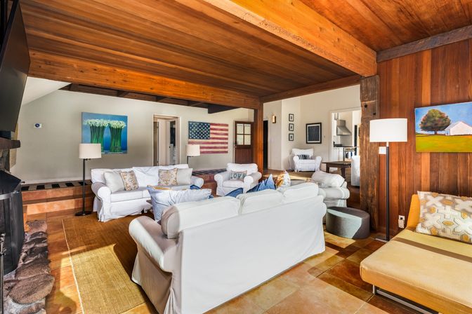 Cozy sunlit living room with exposed wood-beamed ceiling, white slipcovered sofas, stone fireplace, wood-paneled wall, American flag wall art and colorful paintings on warm tile floors.