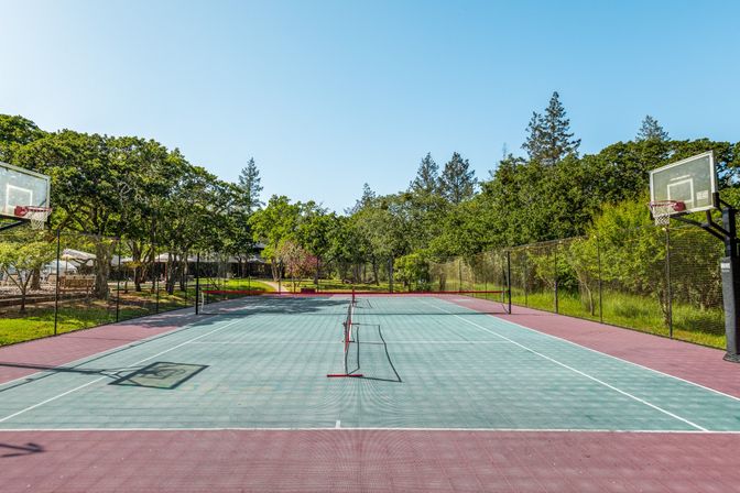 Sunny outdoor multi-sport court with teal surface and red border, portable tennis net down the center and basketball hoops at each end, fenced and surrounded by trees under a clear blue sky