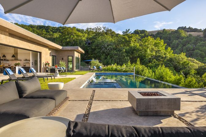 Luxury modern patio with infinity pool and lounge chairs, shaded umbrella, outdoor sofa and concrete fire pit overlooking lush wooded hills, with open-plan living area visible through sliding doors