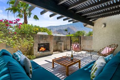 Sunlit mountain-view patio under a wooden pergola with a stone fireplace, teal lounge sofa, wicker chairs with pink cushions, coffee table with wine and glasses, palm trees and bright desert blooms.