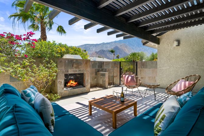 Sunlit mountain-view patio under a wooden pergola with a stone fireplace, teal lounge sofa, wicker chairs with pink cushions, coffee table with wine and glasses, palm trees and bright desert blooms.