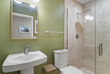 Contemporary bathroom with olive-green accent wall, framed mirror, white pedestal sink and toilet, decorative metal stool, and glass-enclosed beige-tiled shower with rainfall showerhead.