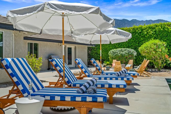 Sunny poolside patio with blue-and-white striped lounge chairs, rolled towels, large white umbrellas and mountain backdrop