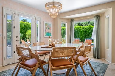 Sunlit dining room with an oval white table, rattan-backed wooden chairs, layered pendant chandelier and patio doors opening to a lush garden and poolside lounge chairs.