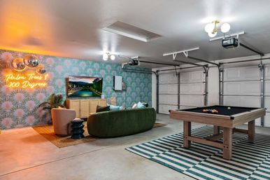 Converted garage game room with pool table on a striped rug, green velvet sofa and armchair, wall TV, palm-print wallpaper, neon "Palm Trees 100 Degrees" sign and mirrored disco balls.