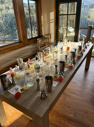 Sunlit wooden table set for a cocktail workshop with shakers, bottles, jiggers and colorful mixers, inside a mountain cabin overlooking pine-covered slopes.