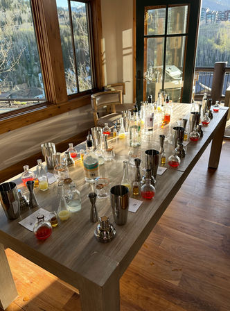 Sunlit wooden table set for a cocktail workshop with shakers, bottles, jiggers and colorful mixers, inside a mountain cabin overlooking pine-covered slopes.