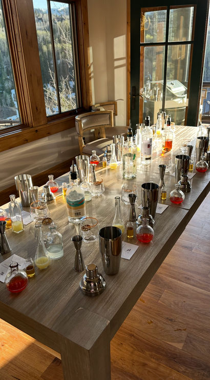 Sunlit wooden table set for a cocktail workshop with shakers, bottles, jiggers and colorful mixers, inside a mountain cabin overlooking pine-covered slopes.
