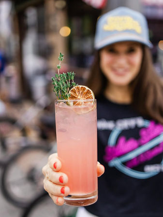 Pink craft cocktail in a tall glass garnished with a dried orange slice and thyme, held forward by a hand with coral nails on an urban outdoor patio.