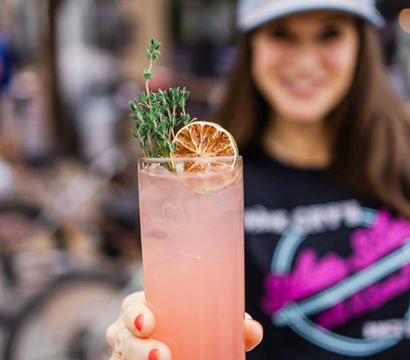 Pink craft cocktail in a tall glass garnished with a dried orange slice and thyme, held forward by a hand with coral nails on an urban outdoor patio.