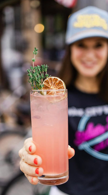 Pink craft cocktail in a tall glass garnished with a dried orange slice and thyme, held forward by a hand with coral nails on an urban outdoor patio.