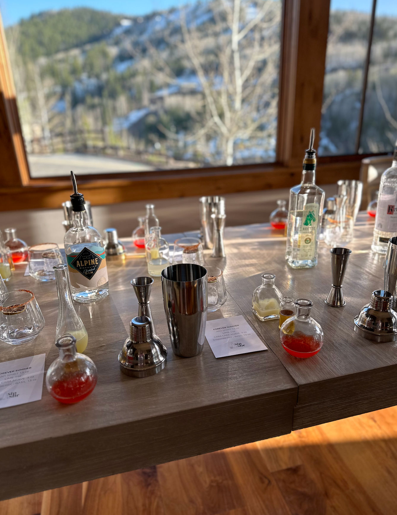 Sunlit mountain-cabin cocktail setup: stainless-steel shaker and jiggers, liquor bottles and small round glass flasks with red and yellow mixers arranged on a wooden table, snowy pine-covered valley visible through a large window.