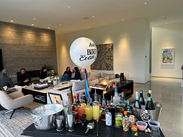 Modern open-plan living room set up for a home birthday party — guests lounging on neutral sofas, a large white birthday balloon, abstract wall art, and a foreground cocktail station with bottles, mixers, ice bucket and garnishes.