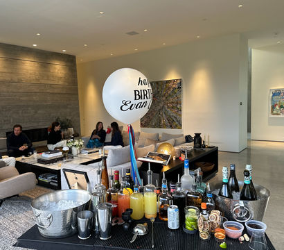 Modern open-plan living room set up for a home birthday party — guests lounging on neutral sofas, a large white birthday balloon, abstract wall art, and a foreground cocktail station with bottles, mixers, ice bucket and garnishes.