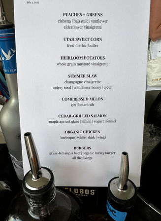 Printed white summer menu dated July 4, 2025 listing dishes like Peaches + Greens, Utah sweet corn, cedar-grilled salmon and burgers, propped among bottles with metal pour spouts.