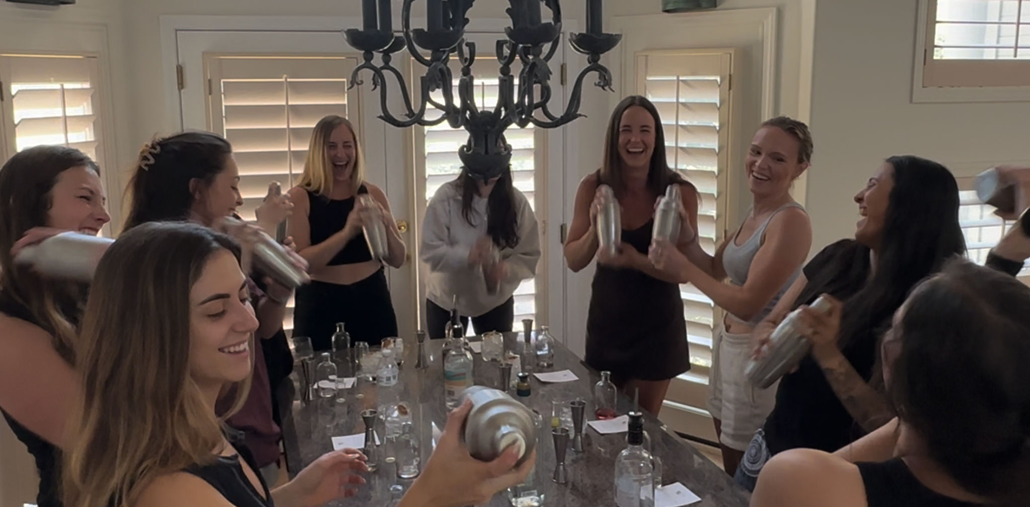 Group of women laughing and shaking cocktail shakers around a kitchen island at a home mixology party, bottles and glassware on the counter under a chandelier and bright window shutters.