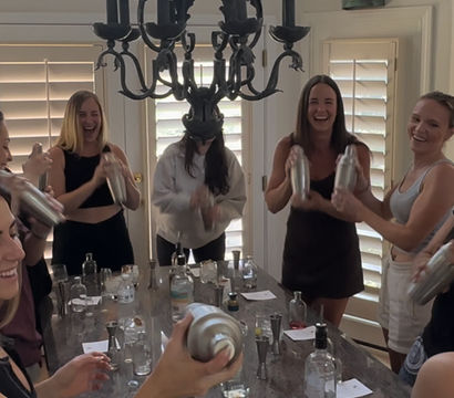 Group of women laughing and shaking cocktail shakers around a kitchen island at a home mixology party, bottles and glassware on the counter under a chandelier and bright window shutters.