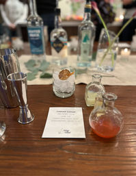 Cocktail mixology setup on a wooden bar: recipe card titled "Forever Summer", ice-filled rocks glass with dried citrus garnish, small glass vials of mixers and blurred spirit bottles in the background.