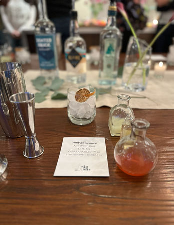 Cocktail mixology setup on a wooden bar: recipe card titled "Forever Summer", ice-filled rocks glass with dried citrus garnish, small glass vials of mixers and blurred spirit bottles in the background.