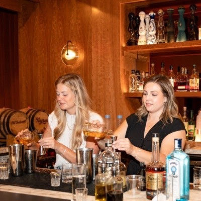Two bartenders mixing craft cocktails at a warm wood-paneled bar, pouring amber spirits into shakers with bottles, glassware and oak barrels displayed on shelves behind them.