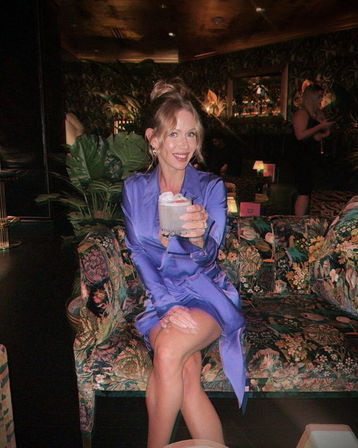 Smiling woman in a purple satin dress seated on a floral sofa in a dim, tropical‑style cocktail lounge, holding a frothy drink toward the camera.