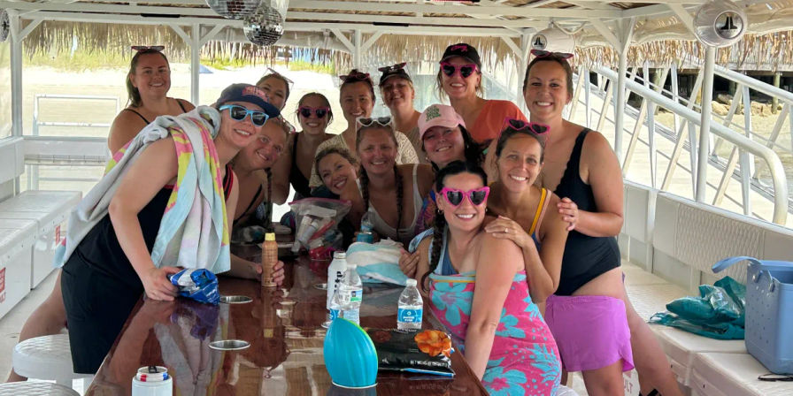 Smiling group of women in colorful swimsuits and sunglasses posing around a wooden table on a covered party boat at a beach dock, towels, drinks, and disco balls overhead
