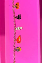Spooky-cute gold chain Halloween charm bracelet with enamel jack-o'-lanterns, witch hat, skeleton and cauldron charms on a bright pink background.