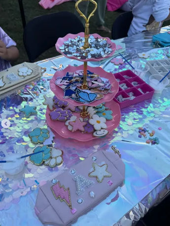 Sparkly outdoor craft table with iridescent sequin tablecloth and a pink three-tier stand holding pastel star, flower and cloud-shaped decorated pieces, rhinestone stickers, small paint cups and an organizer tray.