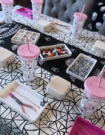 Halloween-themed kids craft table with spiderweb tablecloth, pink ghost tumblers with straws, trays of colorful beads and rhinestones, jewelry pliers and small boxes for DIY bracelets.