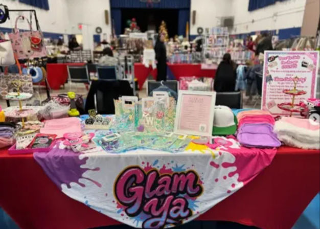 Colorful vendor table at an indoor craft fair displaying beauty and accessory products — holographic bags, pastel pouches, caps, display cards and bright signage on a pink-draped table.