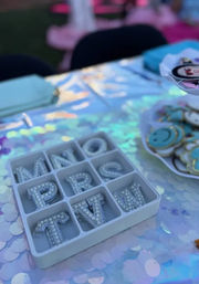 Iridescent party craft table with a white organizer tray of rhinestone alphabet charms on a sequin tablecloth, beside a plate of colorful smiley-face tokens — playful DIY party decor.