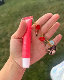 Close-up of a hand holding a bright pink lip-gloss tube with playful red heart, lips and tiny lipstick charms dangling, blurred grassy outdoor background.