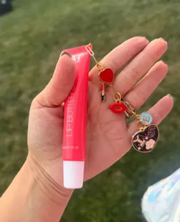 Close-up of a hand holding a bright pink lip-gloss tube with playful red heart, lips and tiny lipstick charms dangling, blurred grassy outdoor background.