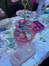 Sparkly outdoor craft table with iridescent sequin tablecloth and a pink three-tier stand holding pastel star, flower and cloud-shaped decorated pieces, rhinestone stickers, small paint cups and an organizer tray.