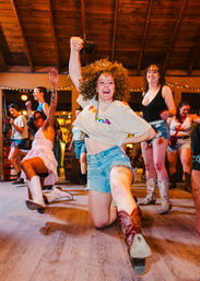 Energetic country-style dance in a rustic indoor hall: woman in denim shorts and cowboy boots lunges forward, curly hair flying, friends and string lights in the background.