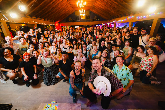 Large diverse group photo at a lively indoor community celebration in a rustic hall with wooden vaulted ceiling, string lights and a chandelier; people smiling and posing on the dance floor with two hosts kneeling front—one holding a white cowboy hat and one wearing a headset.