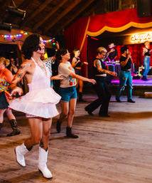 Line dancers at a lively indoor dance hall — a woman in a pink twirl dress and white cowboy boots leads the group in front of a red-curtained stage with string lights and live music.
