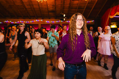 Crowd grooving at an indoor dance party on a wooden-floor hall with colorful string lights and a vaulted wood ceiling, featuring a smiling long-haired person in a purple shirt leading the group.