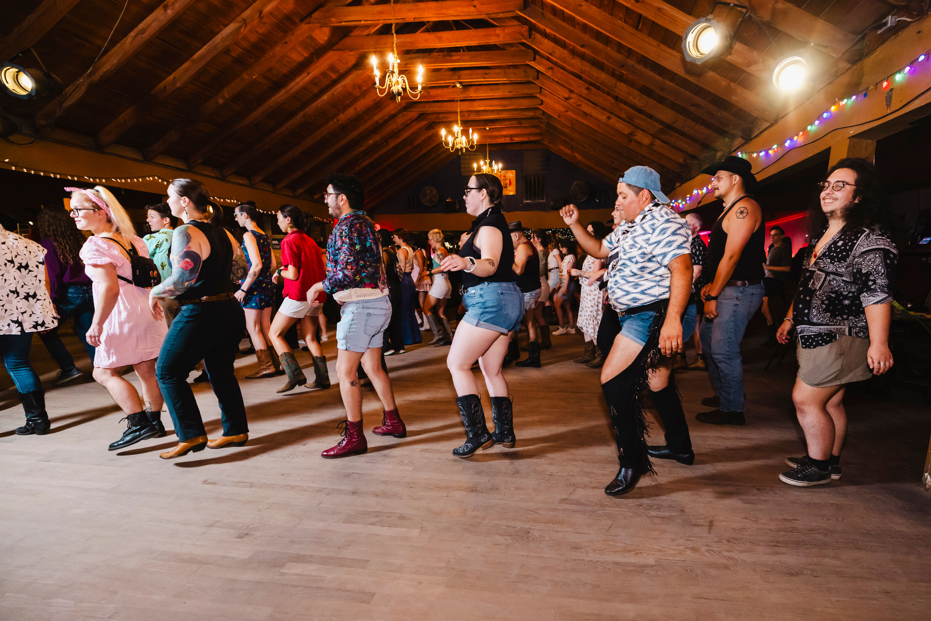 Diverse crowd line dancing in a rustic wooden barn dance hall under chandeliers and string lights, many wearing boots and casual western attire.