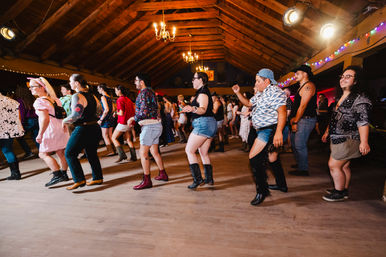 Diverse crowd line dancing in a rustic wooden barn dance hall under chandeliers and string lights, many wearing boots and casual western attire.